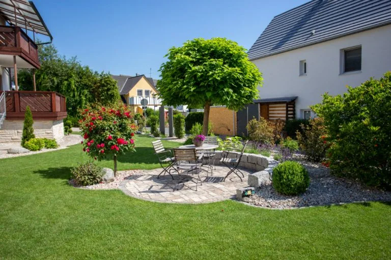 Gartenmöbel auf gepflasterter Terrasse mit Baum und blühenden Sträuchern in gepflegtem Garten.