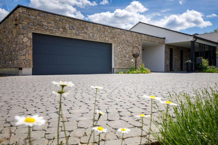 Großflächig gepflasterte Hofeinfahrt mit Natursteinfassade und Betonpflaster in Polygonoptik, Einfahrt pflastern in Burk von BELLA FLORA GmbH.