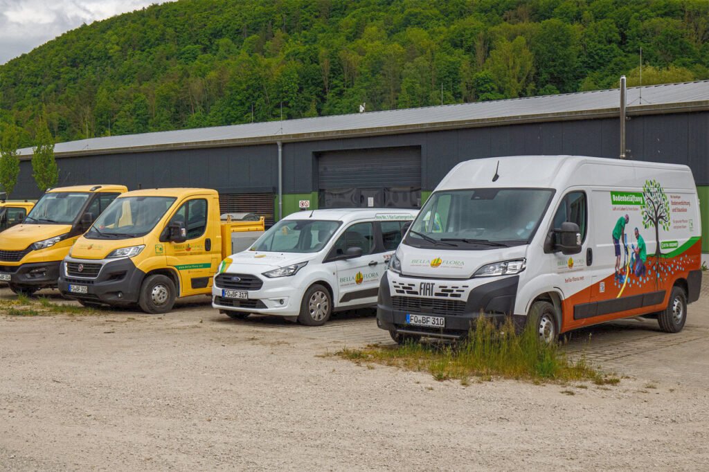 Fuhrpark von BELLA FLORA mit Einsatzfahrzeugen für Garten- und Landschaftsbau auf Betriebshof vor Lagerhalle, professioneller Garten- und Landschaftsbau in Poxdorf von BELLA FLORA GmbH.