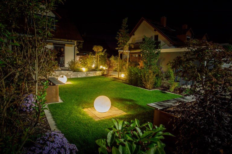 Beleuchteter Garten mit Rasenfläche, Kugelleuchten, Hochbeeten aus Metall, Natursteinmauer und strukturierter Gehölzbepflanzung bei Nacht, Gartenbeleuchtung und Gartengestaltung in Hausen von BELLA FLORA GmbH.