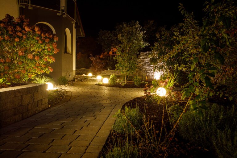 Beleuchteter Gartenweg mit geschwungener Pflasterfläche, Kugelleuchten und strukturierter Stauden- und Gehölzbepflanzung bei Nacht, Pflasterarbeiten und Gartenbeleuchtung in Reuth von BELLA FLORA GmbH.