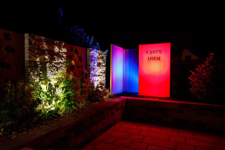 Beleuchtete Sichtschutz- und Gestaltungsmauer mit farbiger Gartenbeleuchtung, Betonsteinmauer und Zierpflanzung bei Nacht, Gartenbeleuchtung und Mauerbau in Streitberg von BELLA FLORA GmbH.