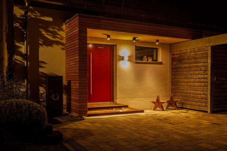 Beleuchteter Hauseingang mit moderner Pflasterfläche, Holzfassade, Stufenanlage und integrierter Außenbeleuchtung bei Nacht, Pflasterarbeiten und Eingangsbereich gestalten in Hirschaid von BELLA FLORA GmbH.