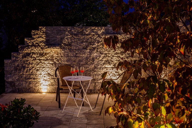 Sitzplatz auf Betonstein-Terrasse mit Bistro-Tisch, Stühlen und beleuchteter Betonsteinmauer bei Nacht, Terrassenbau und Gartenbeleuchtung in Pottenstein von BELLA FLORA GmbH.