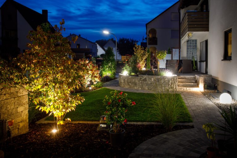 Modern gestalteter Vorgarten mit Rasenfläche, Betonstein-Hochbeet, geschwungenem Pflasterweg, Stauden, Ziergehölzen und professioneller Gartenbeleuchtung bei Nacht, Gartengestaltung und Gartenbeleuchtung in Gößweinstein von BELLA FLORA GmbH.
