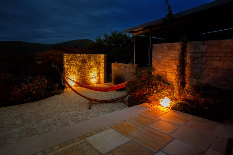 Terrassen- und Gartenbereich mit Beton steinpflaster, Hängematte, massiver Gartenmauer und stimmungsvoller Außenbeleuchtung bei Nacht, Gartengestaltung und Gartenbeleuchtung in Leutenbach von BELLA FLORA GmbH.