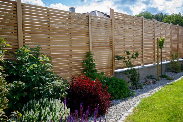 Moderner Holz-Sichtschutzzaun mit horizontalen Lamellen auf Betonsockel, kombiniert mit Staudenbeet, Kiesstreifen und Rasenfläche als Grundstücksabgrenzung, Sichtschutzzaun bauen in Burk von BELLA FLORA GmbH.