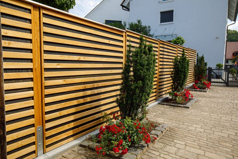 Moderner Holz-Sichtschutzzaun mit horizontalen Lärchenlamellen, dunklem Hintergrund, gepflasterter Einfahrt und strukturierter Bepflanzung, Sichtschutzzaun bauen in Reuth von BELLA FLORA GmbH.