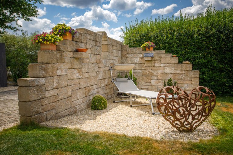 Gestufte Kulissenmauer aus Betonsteinblöcken mit Kiesfläche, Liegebereich und integrierten Pflanznischen als Ruheplatz im Garten, Stützmauer und Gartenbau in Hirschaid von BELLA FLORA GmbH.