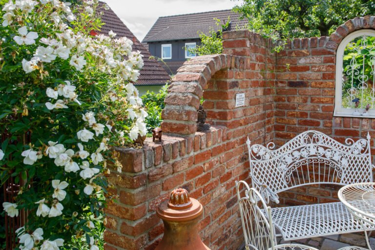 Klinker-Kulissenmauer mit gemauerter Rundbogenöffnung, Sitzplatz und Rosenbewuchs im Hausgarten, Gartenmauer bauen in Kirchehrenbach von BELLA FLORA GmbH.