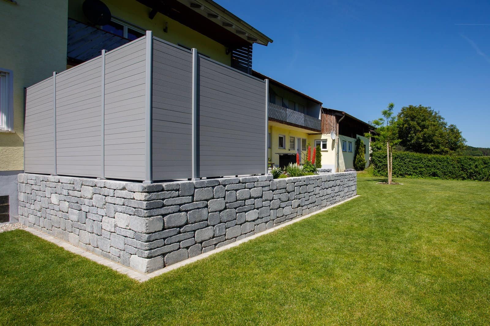 Moderne Sichtschutzanlage aus grauen Paneelen auf massiver Betonsteinmauer mit Rasenfläche im Wohngarten, Sichtschutz bauen in Hausen von BELLA FLORA GmbH.