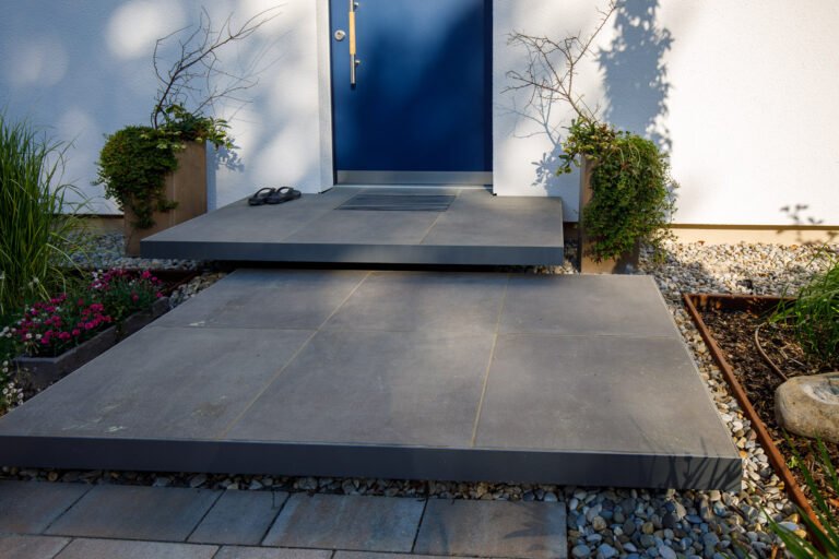 Moderner Hauseingang mit schwebenden Podeststufen aus großformatigen Betonplatten, Kiesbett und symmetrischer Kübelbepflanzung, professioneller Eingangs- und Treppenbau in Neunkirchen am Brand von BELLA FLORA GmbH.