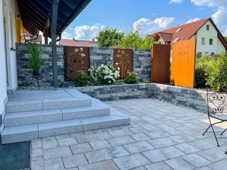 Moderner Terrassenbereich mit Granitblockstufen, gepflasterter Fläche, Naturstein-Stützmauer und Sichtschutzelementen aus Cortenstahl, fachgerechter Terrassen- und Mauerbau in Burk von BELLA FLORA GmbH.