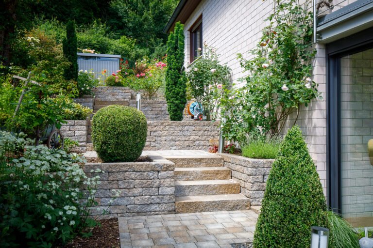 Mehrstufige Gartentreppe aus Betonstein mit seitlichen Stützmauern, gepflastertem Podest und üppiger Bepflanzung entlang der Hausfassade.