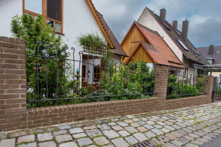 Metallzaun mit Zierelementen auf durchgehender Ziegelmauer vor bepflanztem Vorgarten als hochwertiger Zaunbau in Hirschaid von BELLA FLORA GmbH.