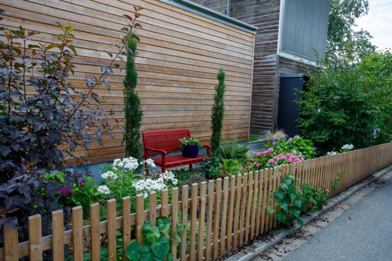 Schmaler Vorgarten mit Holzlattenzaun, Staudenbeet, Ziergehölzen und Sitzbank entlang der Hausfassade, Vorgartengestaltung und Zaunbau in Hausen von BELLA FLORA GmbH.