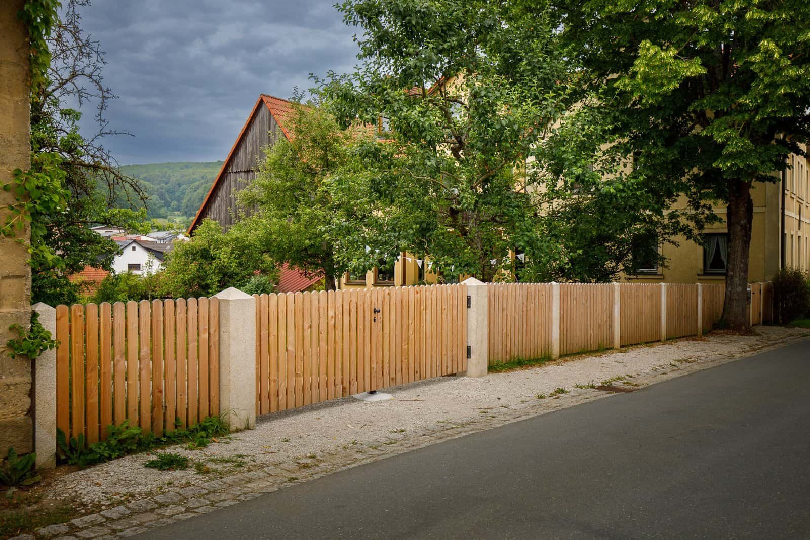 Straßenseitige Holzlatten-Zaunanlage mit Natursteinpfosten und integriertem Gartentor vor Wohnhaus, Zaunbau und Grundstückseinfassung in Eggolsheim von BELLA FLORA GmbH.