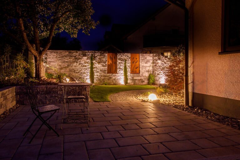 Beleuchtete Terrasse mit Pflasterfläche, Betonsteinmauer und stimmungsvollem Gartenlicht, Pflasterarbeiten und Gartenbeleuchtung in Ebermannstadt von BELLA FLORA GmbH.