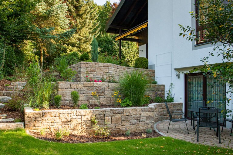 Terrassierter Hanggarten mit Naturstein-Stützmauern, Staudenpflanzung, Ziergräsern und gepflastertem Sitzplatz am Wohnhaus, Garten anlegen in Poxdorf von BELLA FLORA GmbH.