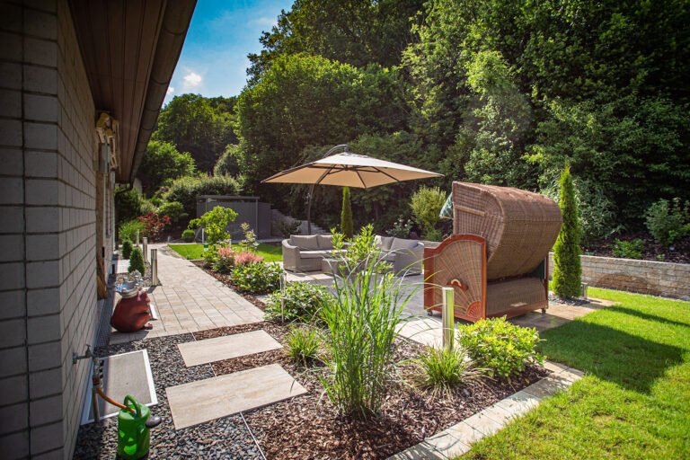 Privatgarten mit gepflastertem Weg, Lounge-Sitzbereich mit Sonnenschirm, Strandkorb, Staudenbeeten und Rasenfläche am Waldrand, Garten anlegen in Hausen von BELLA FLORA GmbH.