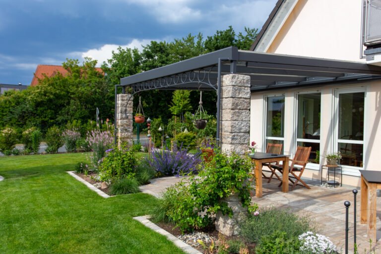 Überdachte Terrasse mit Pergola, Natursteinpfeilern, Staudenbeeten und angrenzender Rasenfläche an einem Einfamilienhaus, Garten anlegen in Reuth von BELLA FLORA GmbH.