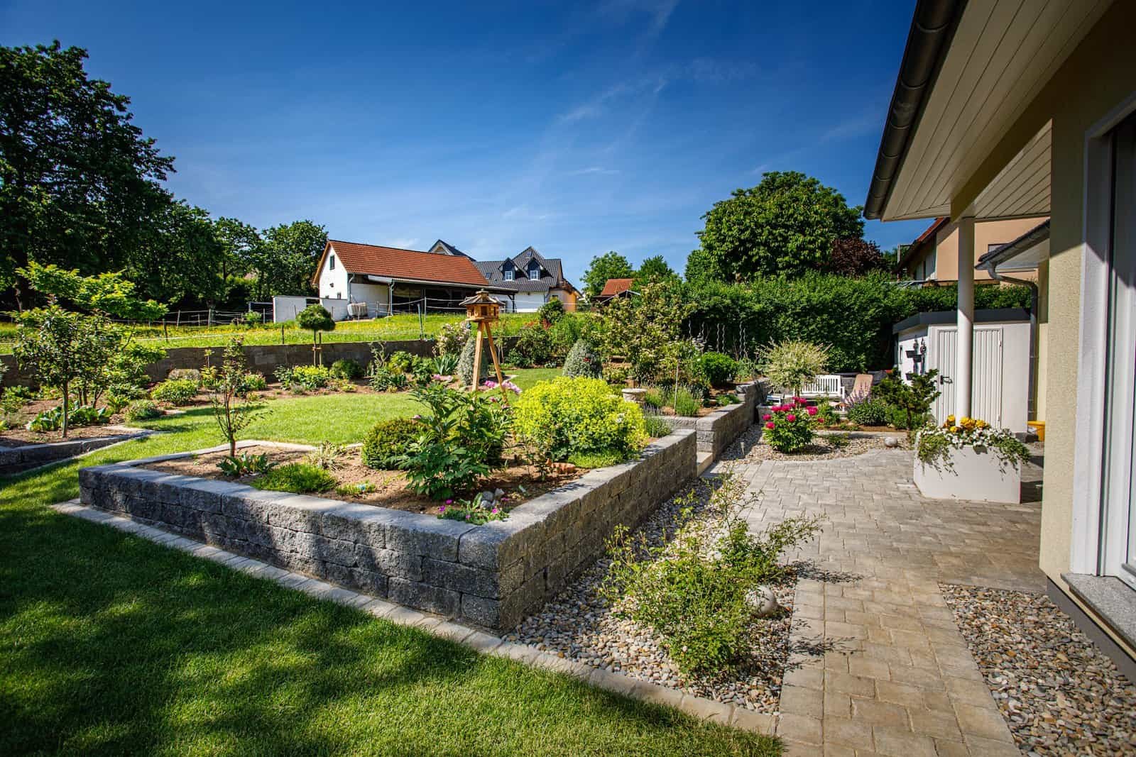 Terrassierter Hausgarten mit Betonstein-Stützmauern, gepflastertem Weg und Staudenbeeten neben einem Einfamilienhaus, Garten anlegen in der Fränkischen Schweiz von BELLA FLORA GmbH.