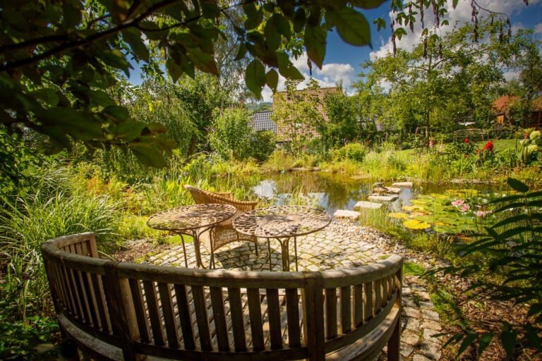 Naturnaher Gartenteich mit Sitzplatz aus Holzbank, Natursteinpflaster, Seerosen und üppiger Uferbepflanzung als professionelle Teich- und Gartengestaltung in Heiligenstadt in Oberfranken von BELLA FLORA GmbH.
