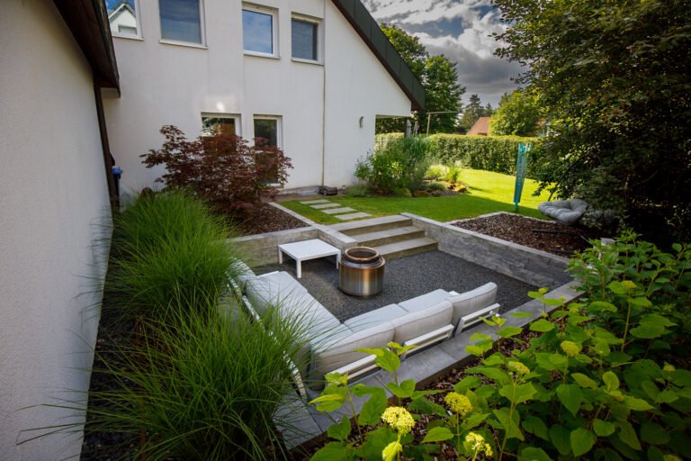 Abgesenkter Sitzplatz mit Betonsteinmauern, integrierter Treppe, Feuerstelle und strukturierter Gartenbepflanzung als hochwertige Gartengestaltung in Pinzberg von BELLA FLORA GmbH.