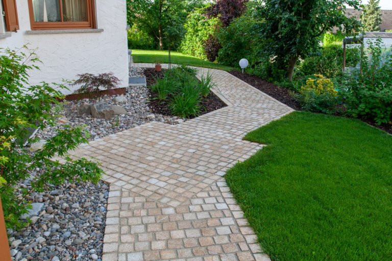 Geschwungener Gartenweg aus hellem Granitpflaster entlang einer Rasenfläche mit angrenzenden Pflanzbeeten an einem Wohnhaus.