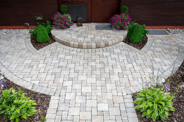Halbrund gepflasterter Eingangsbereich aus hellen Betonsteinen mit Stufe und symmetrischer Bepflanzung vor einem Wohnhaus.