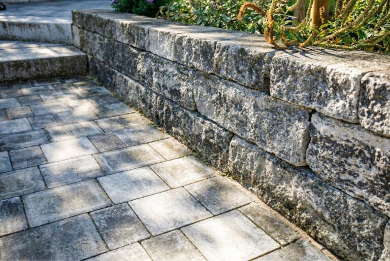 Betonsteinmauer aus grob behauenen Blocksteinen mit angrenzender Betonpflasterfläche – Terrassenbau in Kirchehrenbach von BELLA FLORA GmbH.