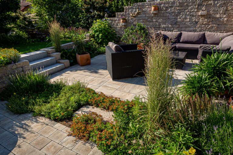 Abgesenkter Sitzplatz mit Betonplatten, Betonsteinmauer, Granitstufen und strukturierter Staudenbepflanzung im Hausgarten, professionelle Garten- und Terrassenanlage in Buckenhofen von BELLA FLORA GmbH.