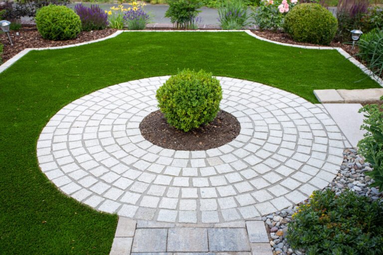 Kreisförmig gepflasterte Natursteinfläche mit mittigem Buchsbaum und angrenzender Rasenfläche, hochwertige Pflaster- und Gartengestaltung in Bamberg von BELLA FLORA GmbH.