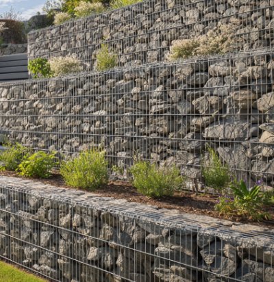 Mehrstufige Gabionen-Stützmauer mit Natursteinfüllung und integrierter Bepflanzung zur Hangabfangung, fachgerechter Mauer- und Erdbau in Weilersbach von BELLA FLORA GmbH.