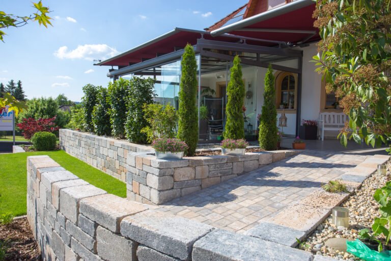 Terrassenanlage mit Betonstein-Stützmauer, gepflasterter Sitzfläche, Wintergarten und strukturierter Bepflanzung zur Gartengestaltung in Weilersbach von BELLA FLORA GmbH.