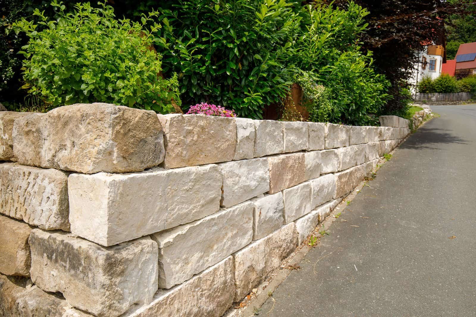 Massive Naturstein-Stützmauer aus Kalkstein entlang einer Straße mit Gehölz- und Staudenbepflanzung, fachgerechter Garten- und Landschaftsbau in der Fränkischen Schweiz von BELLA FLORA GmbH.