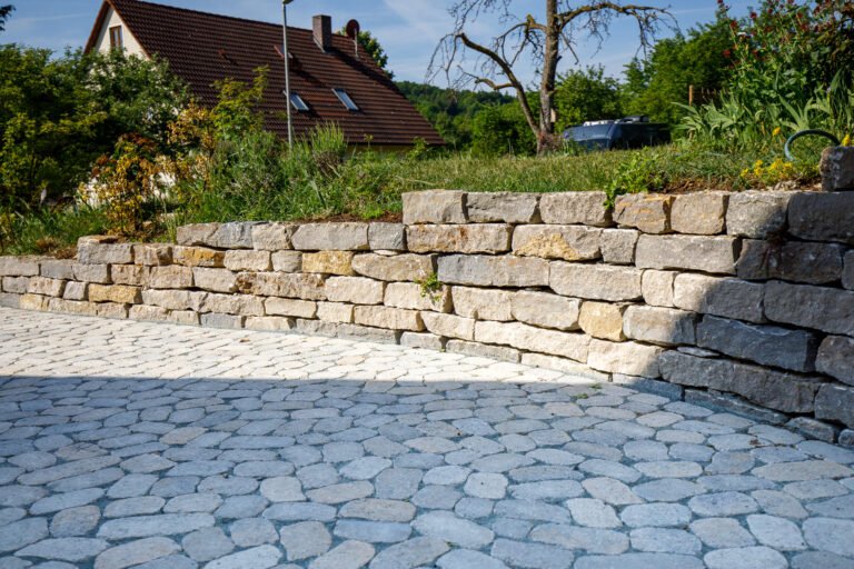 Naturstein-Stützmauer aus Kalkstein mit gepflasterter Einfahrt und Hangabfangung, professioneller Garten- und Landschaftsbau in Forchheim von BELLA FLORA GmbH.