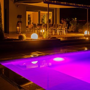 Beleuchteter Pool mit farbiger LED-Unterwasserbeleuchtung, Terrassenbau und Außenbeleuchtung im modernen Garten in Hirschaid von BELLA FLORA GmbH.