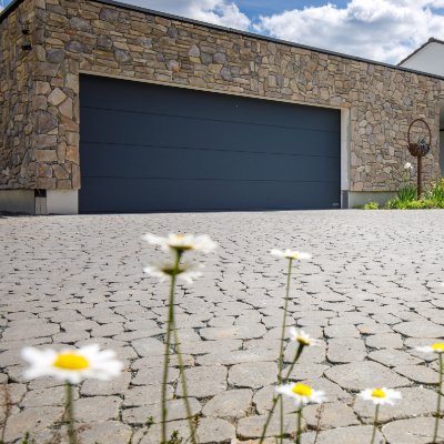 Gepflasterte Hofeinfahrt aus Betonsteinpflaster vor moderner Garage mit Natursteinfassade als hochwertige Pflasterarbeit in Kirchehrenbach von BELLA FLORA GmbH.