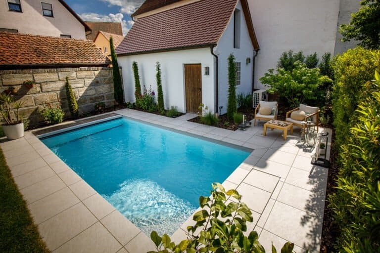 Kompakter Swimmingpool mit Natursteinmauer, heller Terrassenbefestigung und moderner Bepflanzung im Privatgarten in Burk von BELLA FLORA GmbH.