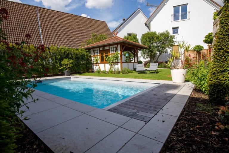 Rechteckiger Swimmingpool mit Holzdeck, großformatigen Terrassenplatten und Gartenpavillon im Privatgarten in Streitberg von BELLA FLORA GmbH.