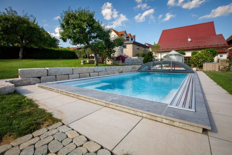 Rechteckiger Swimmingpool mit Schiebeüberdachung, Natursteinmauer und großformatigen Terrassenplatten im Privatgarten in Forchheim von BELLA FLORA GmbH.