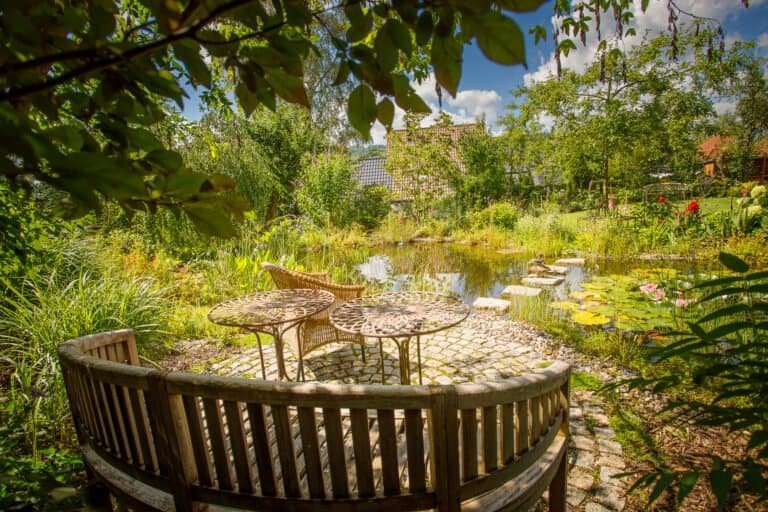 Naturnaher Gartenteich mit Sitzplatz aus Holzbank und Natursteinpflaster als Teichbau und Gartengestaltung in Effeltrich von BELLA FLORA GmbH.