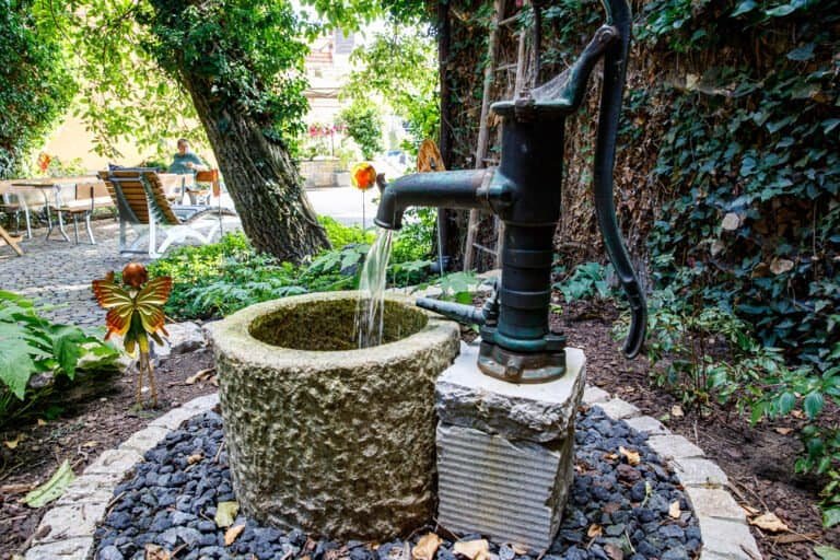 Historische Schwengelpumpe mit Natursteinbrunnen und Kiesbett als Wasserelement im Gartenbau in Leutenbach von BELLA FLORA GmbH.