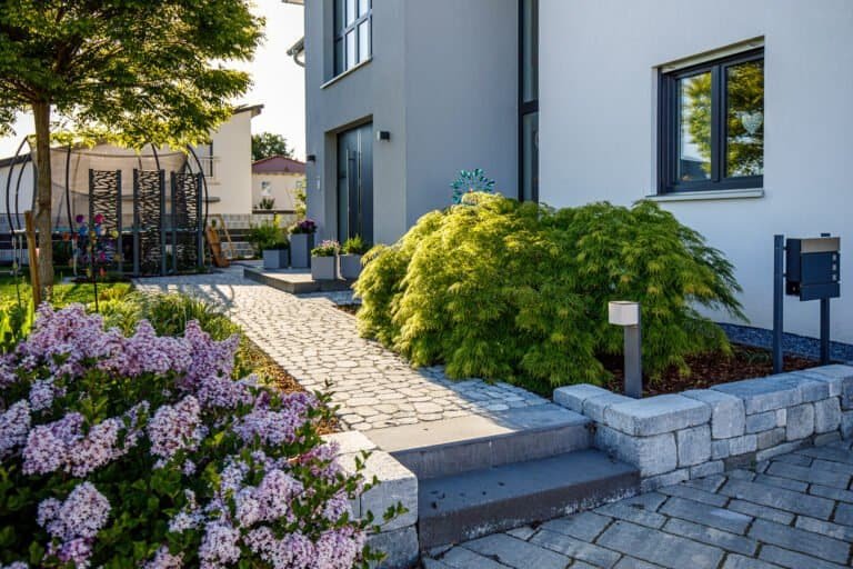 Moderner gepflasterter Eingangsweg mit Betonsteinmauer, Ziergehölzen und Beleuchtung im Vorgarten + Pflasterarbeiten und Gartengestaltung + Buttenheim von BELLA FLORA GmbH.