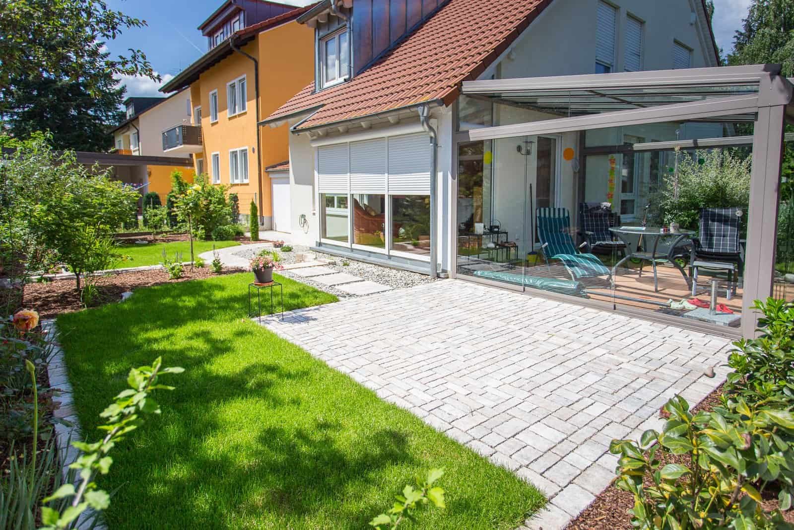 Modern angelegte Terrasse mit Betonpflaster, Glasüberdachung und Rasenfläche – Terrassenbau in Kirchehrenbach von BELLA FLORA GmbH.