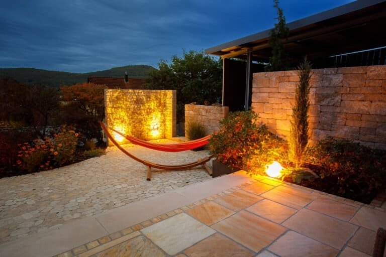 Modern gestaltete Naturstein-Terrasse mit beleuchteter Betonsteinmauer und Hängematte am Abend – Terrassenbau in Hirschaid von BELLA FLORA GmbH.